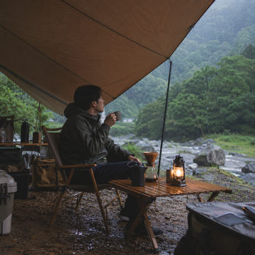 雨天露營時，露營者坐在天幕下喝咖啡，雨水順著天幕邊緣流下，營地呈現濕潤但舒適的氛圍。